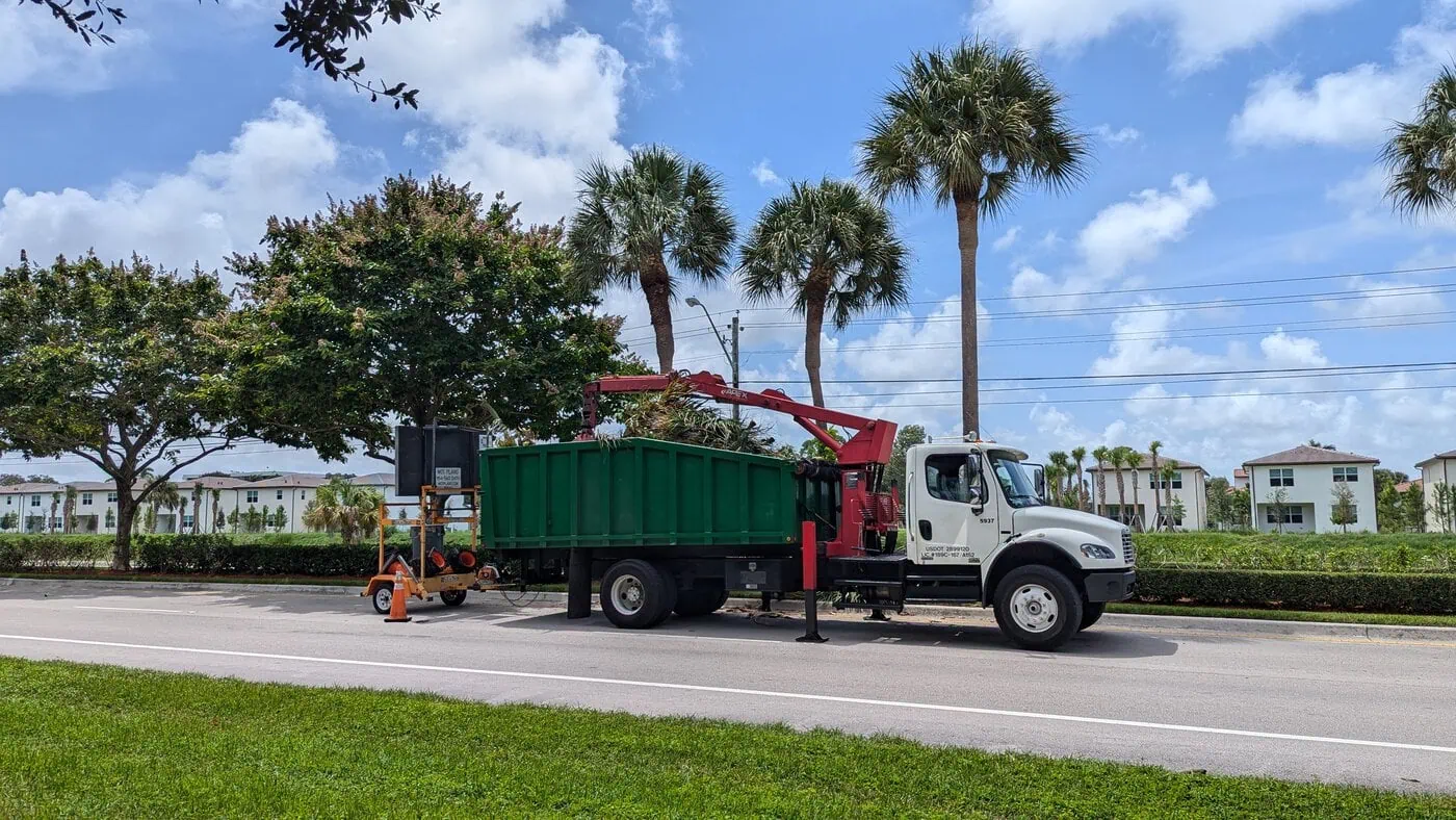 A white utility truck with a green dump bed is parked on a street under palm trees. A crane arm is loading debris into the truck. Nearby is an electronic road sign. Buildings and blue sky with clouds are in the background.